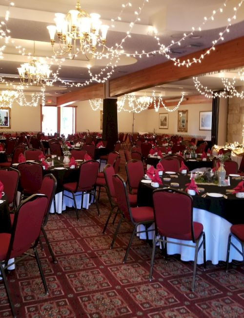 A decorated banquet hall with tables set for an event, featuring red chairs, white tablecloths, and string lights hanging from the ceiling.