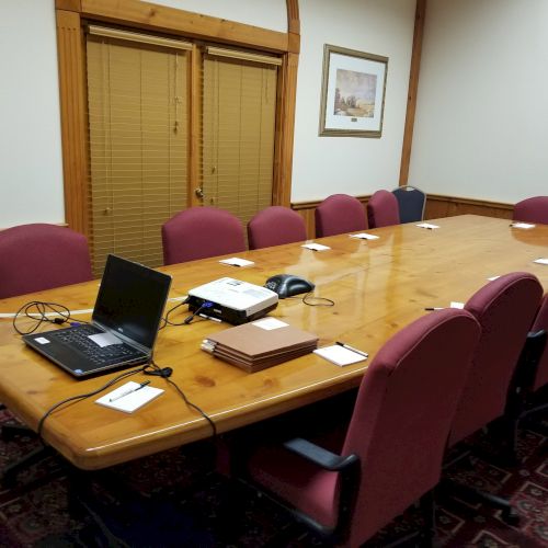 A conference room with a large wooden table, red chairs, a laptop, phone, notepads, and framed art on the walls.