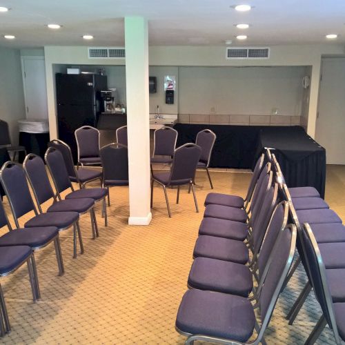 The image shows an empty conference room with rows of chairs arranged in a U-shape, beige carpeting, and a small stage at the front.