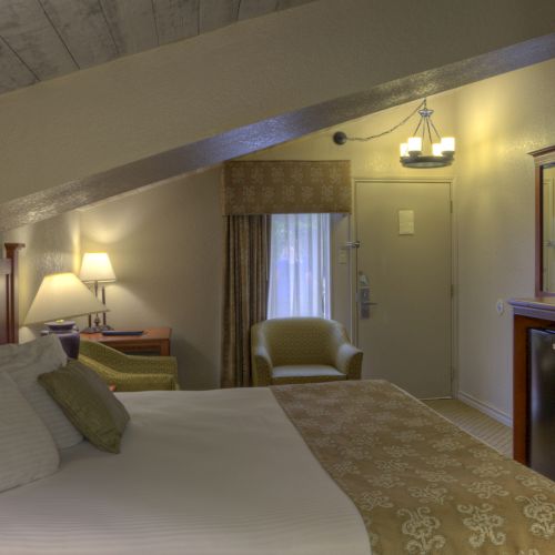 A cozy hotel room with a bed, TV, armchair, and lamp. Warm lighting and slanted ceiling create a comfortable atmosphere.