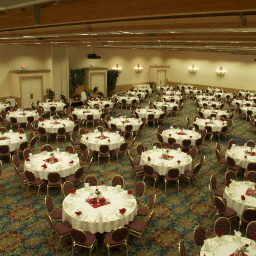 The image shows a large banquet hall with numerous round tables covered with white tablecloths and set with chairs, ready for an event.