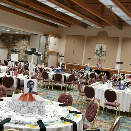 The image shows a banquet hall with round tables set for an event, featuring floral centerpieces and patterned chairs.