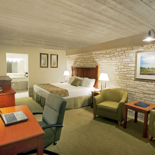 A cozy hotel room with a bed, desk, armchairs, and a painting on the wall; a laptop is on the desk, and lamps light the space.