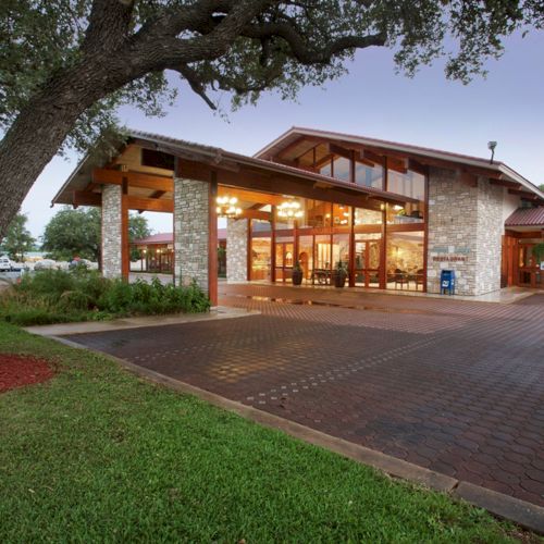 The image shows a modern building with stone walls, large windows, and a gabled roof, set among green landscaping and trees.