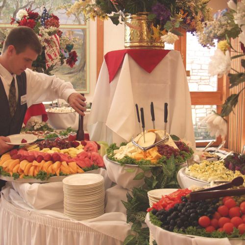 A waiter sets up a buffet with various fruits and cheeses on elegantly arranged tables with floral decor.