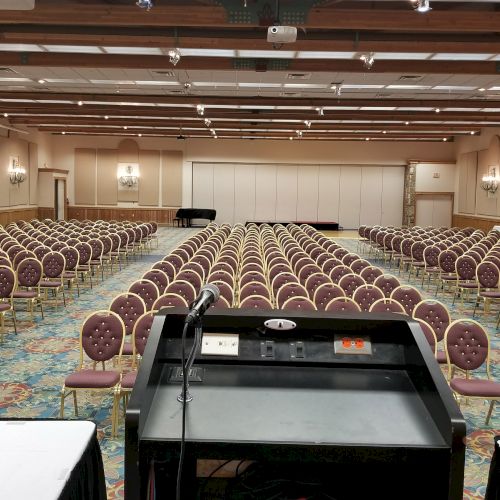 The image shows an empty conference room with rows of chairs facing a podium and tables, prepared for an event or presentation.