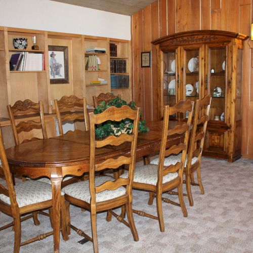 The image shows a dining room with a wooden table set, chairs, a hutch with china, bookshelves, and wood-paneled walls.