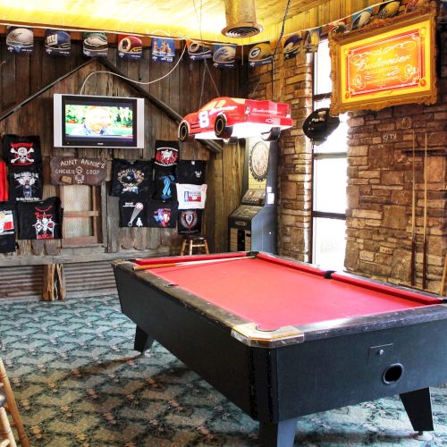 A game room with a red-felt pool table, bar stools, a TV, sports memorabilia, and a rustic decor style.