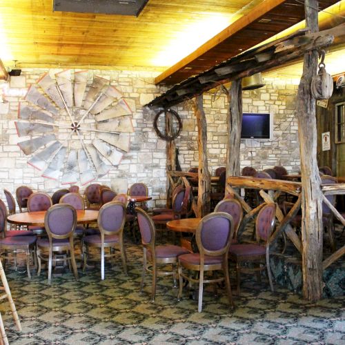 A rustic restaurant interior with wooden beams, round tables, and chairs, featuring stone walls and a patterned floor, creating a cozy ambiance.