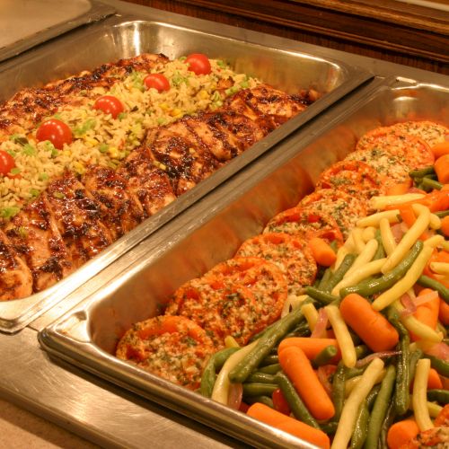 A buffet spread featuring a dish with rice, cherry tomatoes, chicken, and another with vegetables like carrots, green beans, and breaded patties.