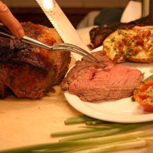 A person is slicing a roast beef with a knife, placing slices on a plate with stuffed potato, tomato, and vegetables.