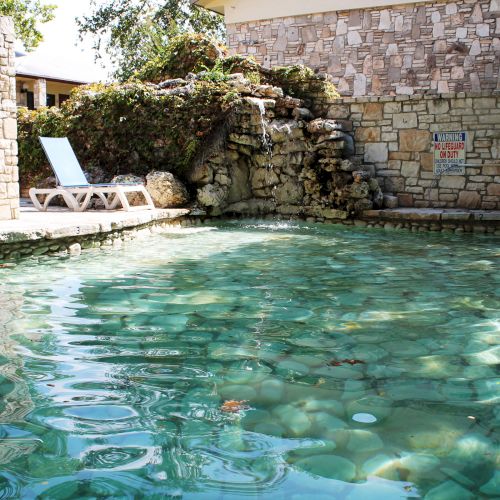 A stone-walled pool with clear water and lounge chairs beside it, under a partially shaded area with trees around.