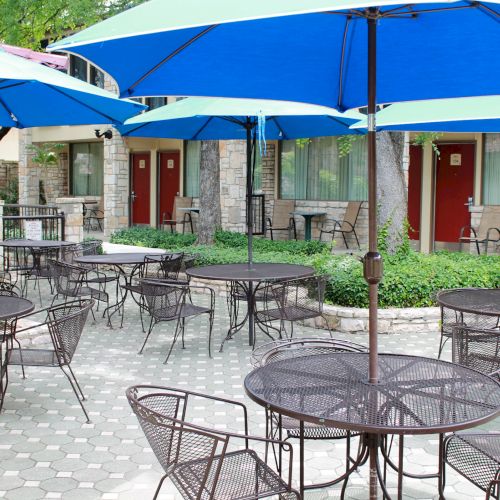 Outdoor patio with metal tables, chairs, and blue umbrellas. Stone building and red doors in the background complete the scene.