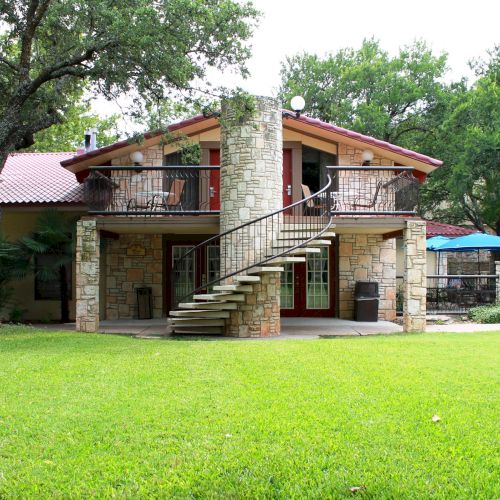 A two-story stone house with a spiral staircase leading to a balcony, surrounded by trees and a grassy lawn.