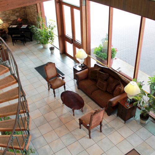 The image shows a cozy room with a spiral staircase, large windows, a sofa, chairs, tables, and potted plants, creating a welcoming atmosphere.