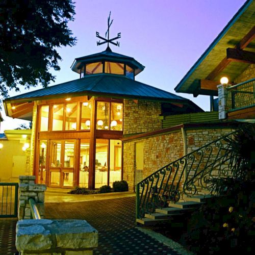 The image shows a well-lit, modern building with large windows and stone architecture at twilight, featuring a winding staircase.