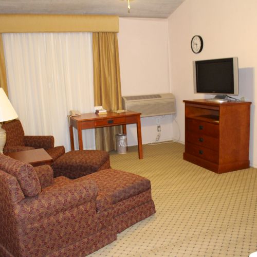 A hotel room with two chairs, a lamp, a TV on a wooden stand, and a small desk near a curtained window.