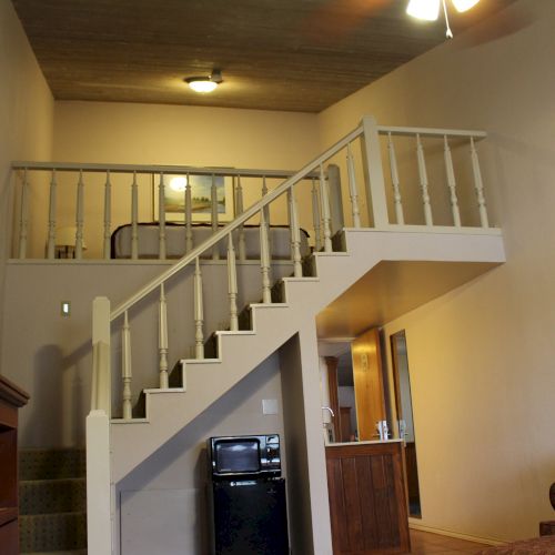 The image shows a loft-style room with a staircase leading to an upper bed area, and a small kitchen area below.