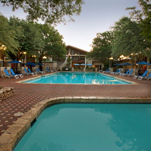 The image shows two swimming pools surrounded by lounge chairs and umbrellas, set within a courtyard with trees and a building in the background.
