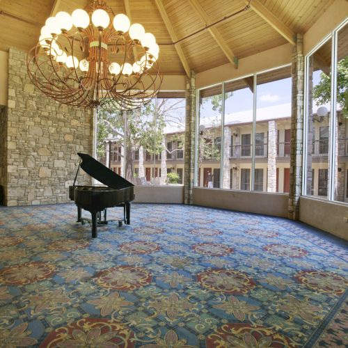 A grand piano sits on a patterned carpet in a spacious room with large windows and a chandelier, overlooking a courtyard.