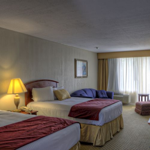 The image shows a hotel room with two beds, a lamp, and a window with curtains letting in natural light.