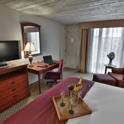 A hotel room with a bed, TV, desk, chair, sofa, lamp, and a tray with drinks, overlooking a window with curtains drawn.