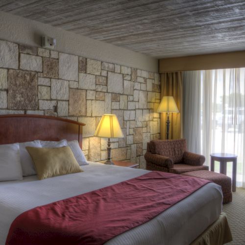A cozy hotel room with a large bed, decorative stone wall, two lamps, a chair, and a large window with curtains.