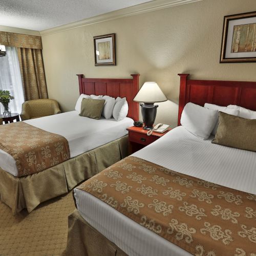 The image shows a hotel room with two double beds, elegant decor, a lamp, chairs, and framed art, creating a cozy and inviting atmosphere.