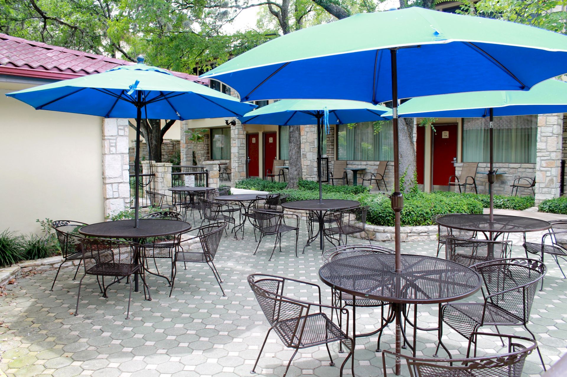 The image shows an outdoor patio area with metal tables and chairs under large blue umbrellas, surrounded by buildings and trees.