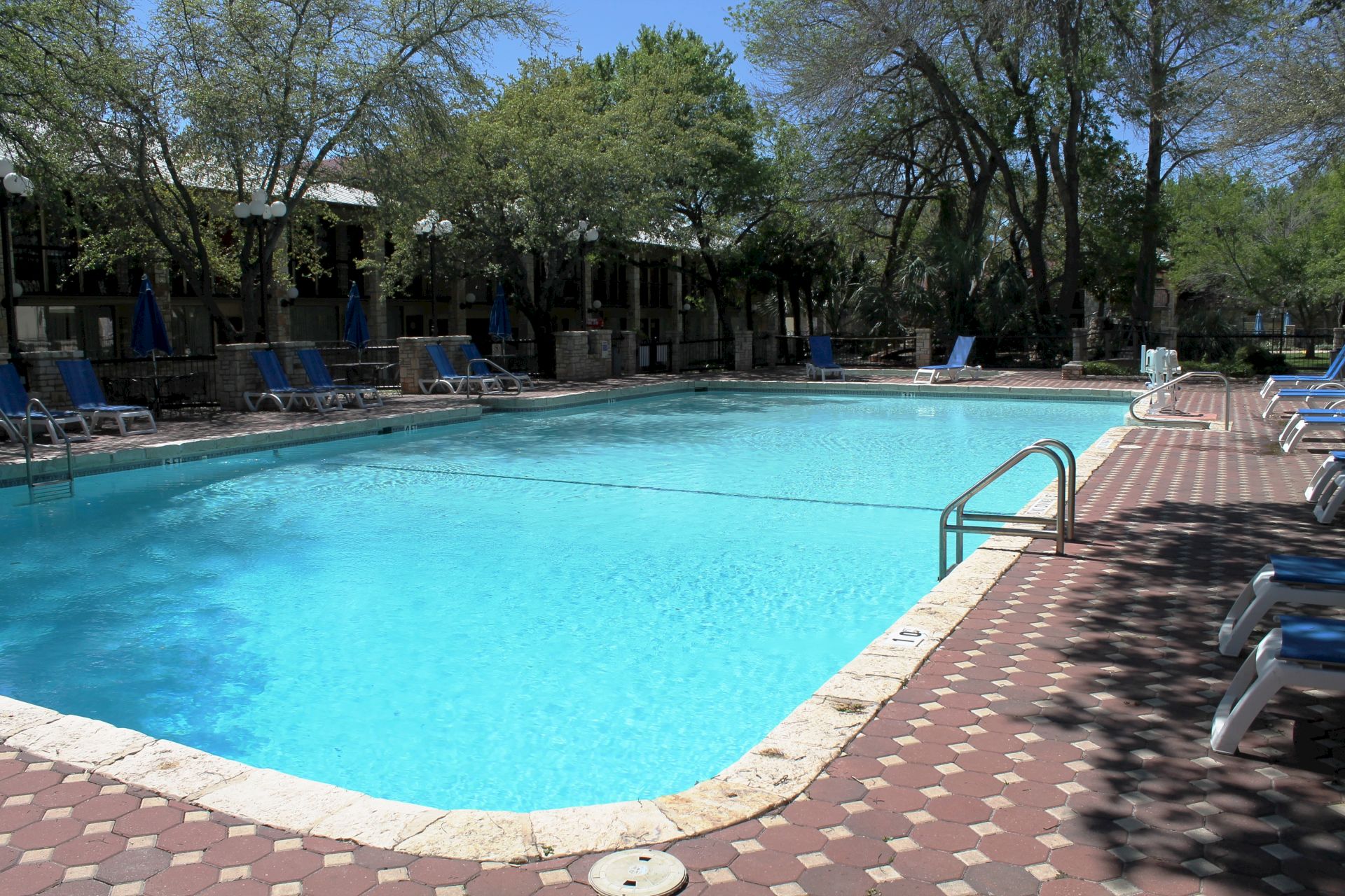 An outdoor swimming pool with clear blue water, lounge chairs, and umbrellas, surrounded by trees and stone walls.