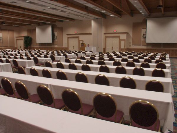 A large conference room with rows of chairs and tables, white tablecloths, and projection screens at the front.
