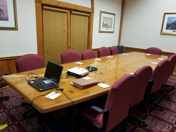 A conference room with a long wooden table, red chairs, a laptop, a phone, notepads, and a lined carpet. Walls have framed artwork.