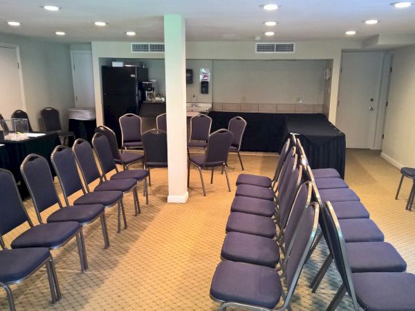 A conference room with rows of blue cushioned chairs arranged theater-style, a long table at the back, neutral walls, and a central pillar.