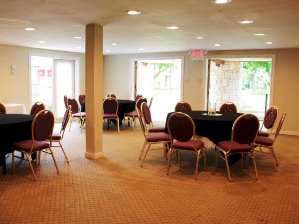 The image shows an empty room set up with round tables, each surrounded by red chairs, and natural light coming through large windows.