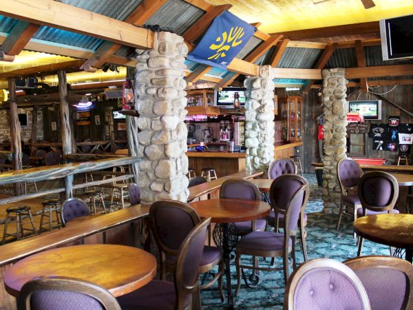 The image shows an empty restaurant or bar with wooden tables, cushioned chairs, stone pillars, and a rustic interior design.