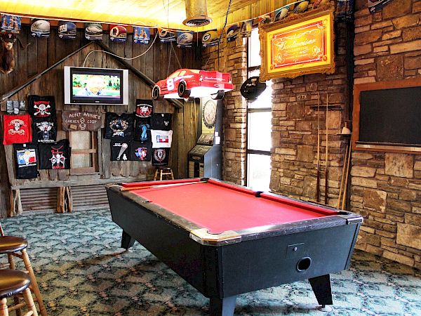 A rustic game room with a red pool table, bar stools, sports decor, and a mounted TV, creating a cozy atmosphere.