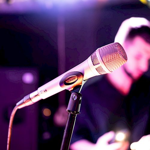 A close-up of a microphone on a stand with a blurred musician performing in the background, in a dim, club-like setting.