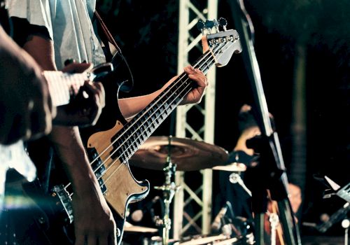 A close-up of a live band playing: guitarist&rsquo;s hands on a bass guitar, drum kit in the background, stage lighting and equipment visible, performing on stage.