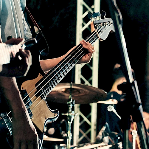 A close-up of a live band playing: guitarist&rsquo;s hands on a bass guitar, drum kit in the background, stage lighting and equipment visible, performing on stage.