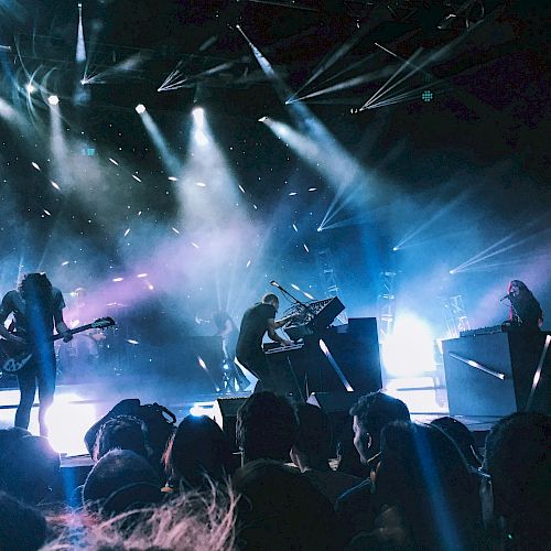 A live concert with a band performing on stage, bright spotlights, and an audience watching intently.