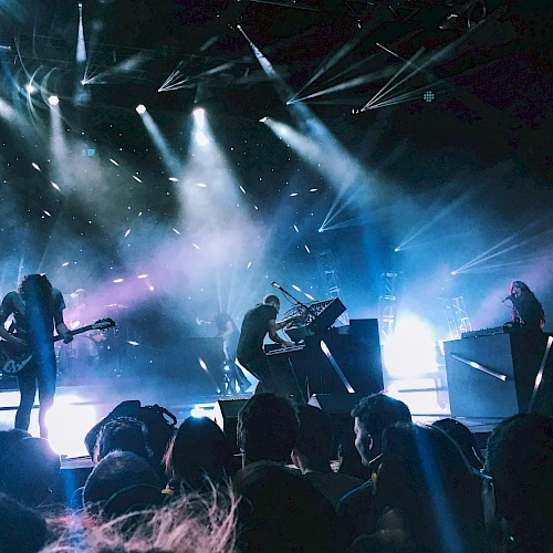 A live concert with a band performing on stage, bright spotlights, and an audience watching intently.