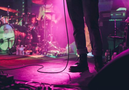 A concert stage with a singer&rsquo;s legs and microphone cord, purple lighting, drums, and gear in the background, live music vibe at night.