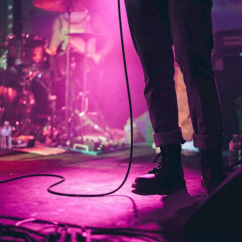 A concert stage with a singer&rsquo;s legs and microphone cord, purple lighting, drums, and gear in the background, live music vibe at night.