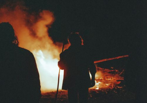 Two people stand near a large bonfire at night, silhouettes against orange flames and smoke, creating a dramatic, fiery scene.