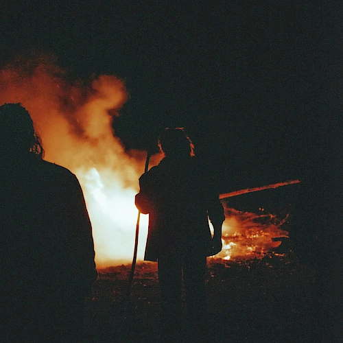 Two people stand near a large bonfire at night, silhouettes against orange flames and smoke, creating a dramatic, fiery scene.