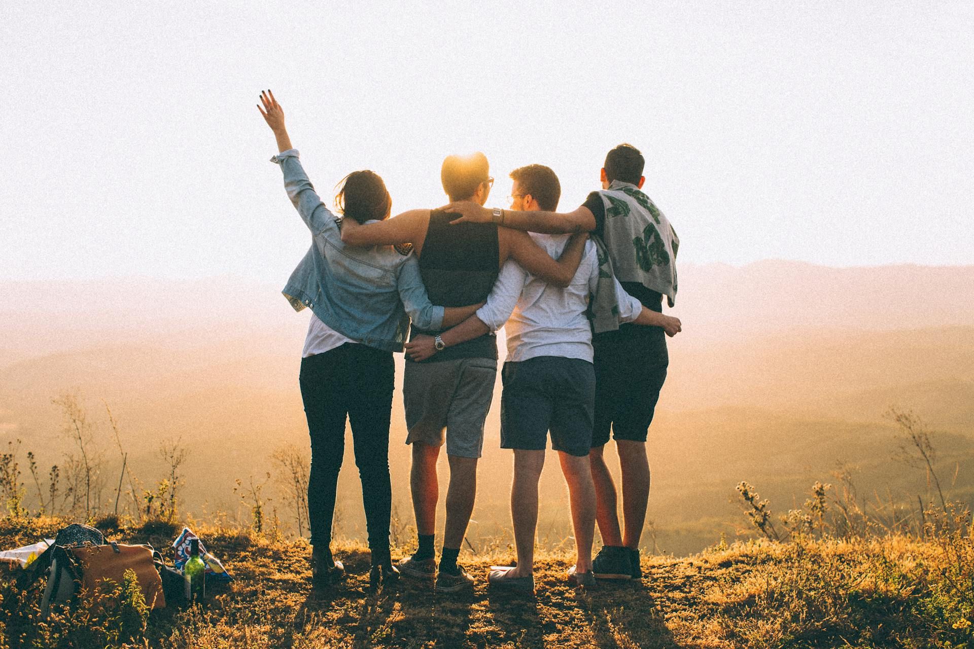 Group of friends in a desert landscape hug and raise arms toward the sky at sunset.