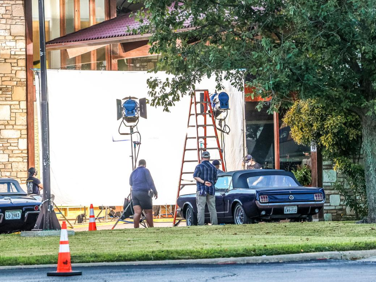 Construction crew setting up lights on a white-wall set with ladders, trucks, cones, and workers near a tree and building.