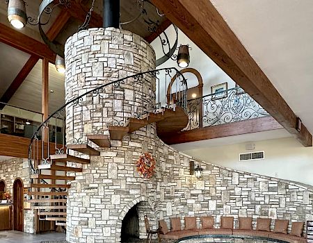 A stone spiral staircase winds around a tall, brick oven feature inside a rustic, vaulted room with wooden beams and tiled floor.