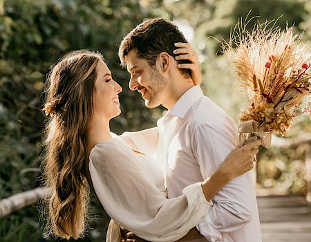 A smiling couple in white clothes share a tender moment, holding a bouquet of dried wheat and flowers outdoors.