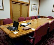 A conference room with a long wooden table, laptops and papers, purple chairs, blinds, and framed landscape paintings on the walls.